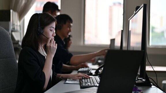 年轻的亚洲呼叫中心女接线员戴着耳机在电脑上工作