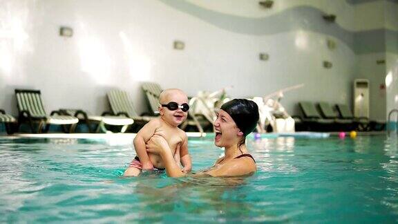 年轻的母亲抱着戴着防护眼镜的小男孩在游泳池里教他如何游泳宝宝用脚在水里踢开始在室内游泳池游泳