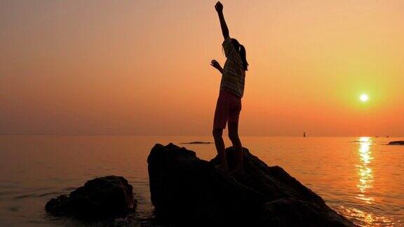 女孩的剪影站在岩石上举起双手在早上日出的海滩上生活理念