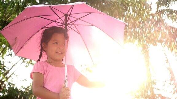 慢动作的可爱的亚洲小女孩拿着伞并有乐趣与雨和阳光玩