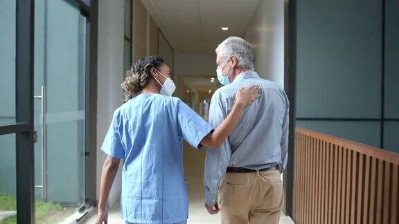 非裔美国女医生走在医院走廊上旁边是一位老年患者两人在COVID-19大流行期间都戴着防护口罩