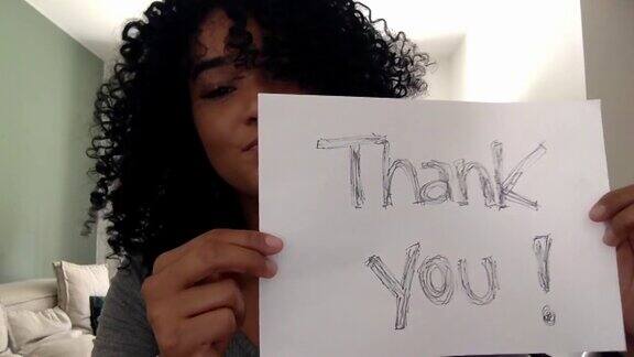 年轻女子拿着写有“谢谢你”的纸在家-摄像头的观点