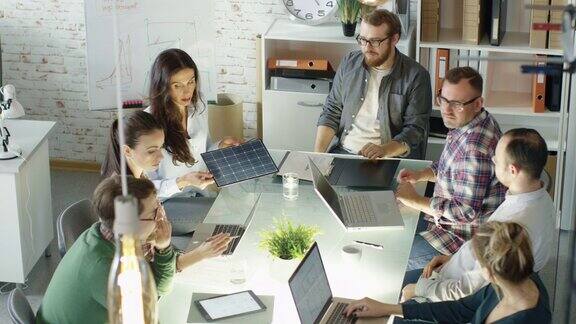 一群创意青年举行太阳能电池板技术战略规划会议他们正坐在明亮而现代的阁楼办公室里的大玻璃桌子旁桌上放着打开的笔记本电脑