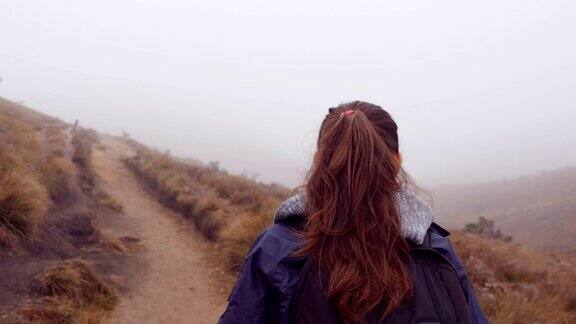 登山妇女在潮湿多雾的天气里穿着雨衣背着背包在山上行走年轻女孩在旅行途中走山路跟着女游客走在路上后视图特写