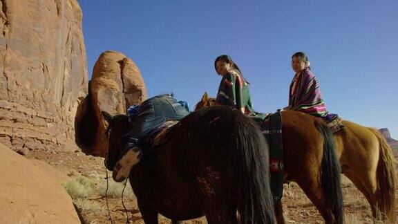 一个十岁的美国土著(纳瓦霍)男孩从岩石爬到他的马而两个十几岁的女孩在亚利桑那州犹他州纪念碑谷沙漠的景观旁边的一个大的岩石形成在一个晴朗明亮的日子