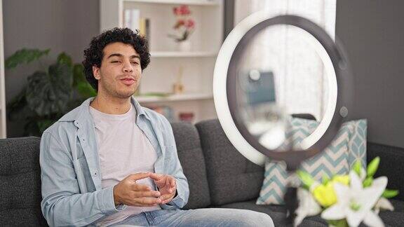 年轻的拉丁男子坐在沙发上录制视频教程在家里