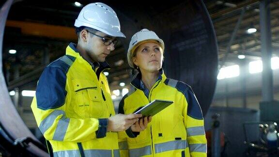 男和女工业工程师在安全帽讨论新项目同时使用平板电脑他们在做计算工程决策他们在重工业制造厂工作