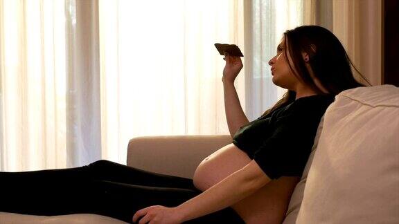 年轻孕妇躺在沙发上一边吃着巧克力一边看电视