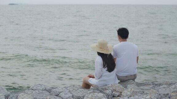 男人和女人夫妇坐在海边的石头上享受假期夏天愉快地说着笑着在晚上看海滩上的景色