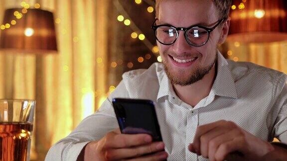 帅气的胡子男子一边在酒吧里喝啤酒一边用智能手机微笑