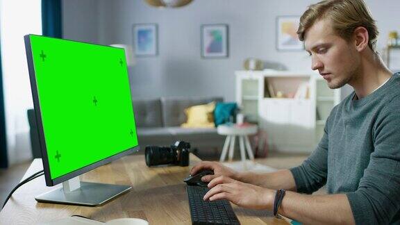 年轻帅气的摄影师在一个绿色模拟屏幕个人电脑上工作而坐在他的书桌在舒适的公寓