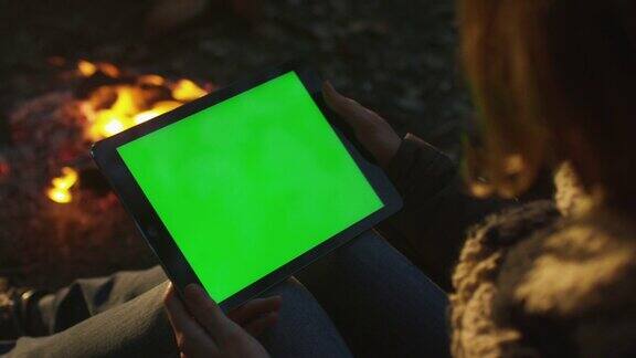 晚上女孩在篝火旁拿着一台绿色屏幕的平板电脑