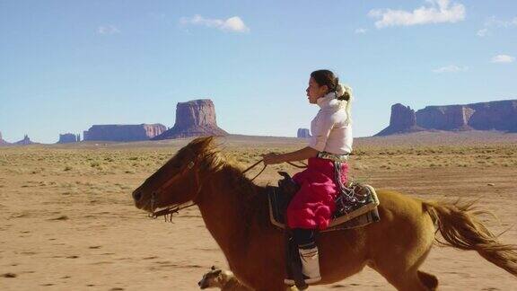 在一个晴朗晴朗的日子里一个十几岁的印第安女孩穿着传统的纳瓦霍服装骑着她的马疾驰在纪念碑谷沙漠和她的宠物狗