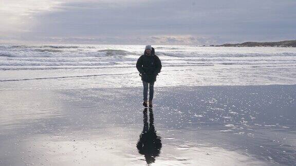 一名男子沿着阳光明媚的潮湿海滩走向摄像机