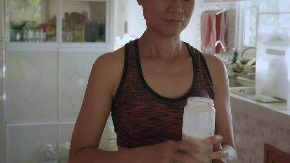 穿着运动服的亚洲年轻女子用蛋白粉制作奶昔在家里的厨房里喝蛋白奶昔