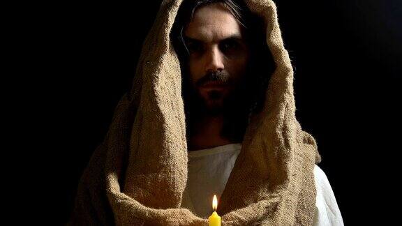 耶稣基督穿着长袍拿着燃烧的蜡烛在黑暗的背景下光明