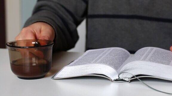 一个男人读着《圣经》翻着书页从杯子里喝水