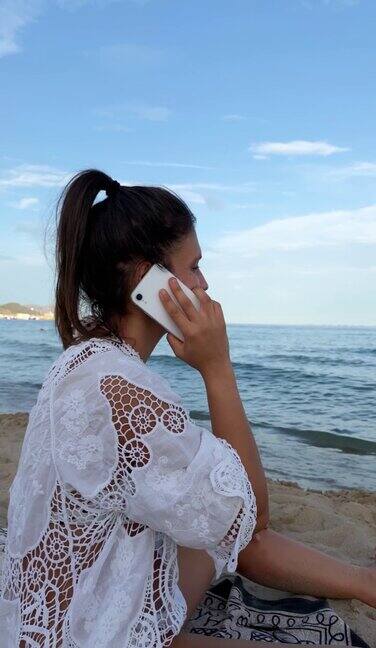 身穿白色沙滩裙的褐发女子坐在海边的毛巾上打电话慢动作垂直视频的年轻女孩使用她的智能手机