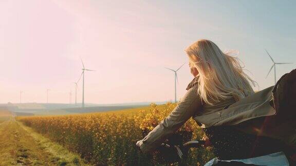 女人骑着自行车沿着油菜籽田走远处有风力发电机