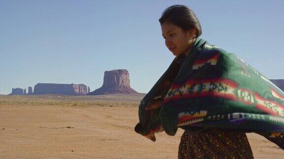 一个十几岁的美国土著女孩在一个晴朗、明亮的日子里在纪念碑谷沙漠与巨大的岩层之间用传统的纳瓦霍毯子裹着她的肩膀的慢动作拍摄
