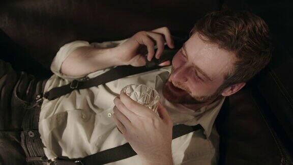 一个喝醉酒的男人下班后躺在沙发上一边喝着威士忌一边微笑着打电话