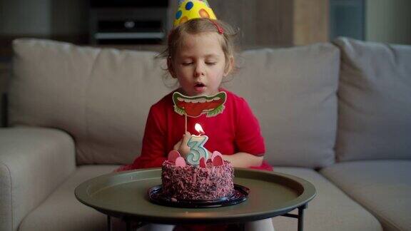 幼儿园女孩戴着生日帽独自在家吹灭巧克力蛋糕上的3号蜡烛孩子独自在家庆祝生日
