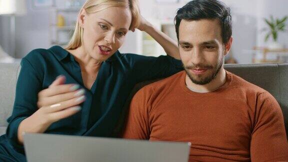 快乐的女朋友和男朋友坐在家里的沙发上用笔记本电脑浏览互联网做电子购物使用流媒体服务幸福家庭在舒适的客厅