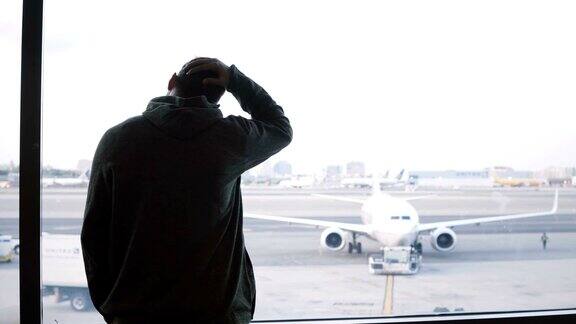 肩扛大包的男性旅客走到机场候机厅窗口欣赏飞机和汽车的景色