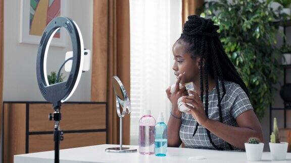 美丽的非洲女性博主展示如何使用化妆品在镜子和照相手机前她在家实时录制视频面部皮肤护理涂抹保湿霜