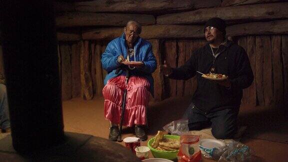 一个40多岁的美国土著男子(纳瓦霍人)跪在霍根(纳瓦霍人)小屋的地板上一边吃着食物一边与他人交谈他坐在旁边的一位老年妇女也在吃东西