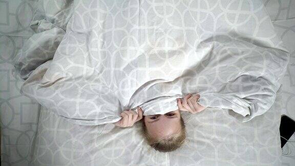 女孩微笑着躲在毯子下面躺在床上看着相机