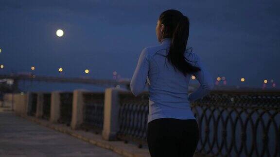 晚上慢跑在一个夜城的背景下一个穿着运动服的女孩沿着空旷的夜堤跑着后视图