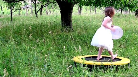一个可爱的女孩在自然花园里跳舞小女孩在小蹦床上又跳又跳小女孩穿着白色的婚纱