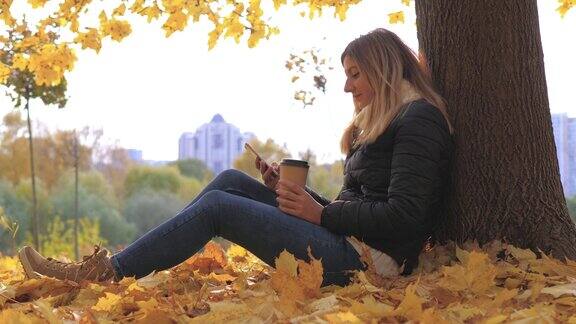 一个女人坐在枯黄的落叶树旁一边用手机应用一边喝咖啡