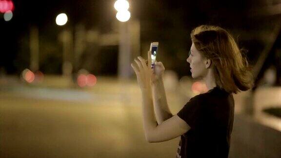 美丽的年轻女子在夜晚的街道上用智能手机拍照