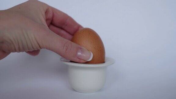用手将棕色鸡蛋放入白色背景的白色蛋杯中