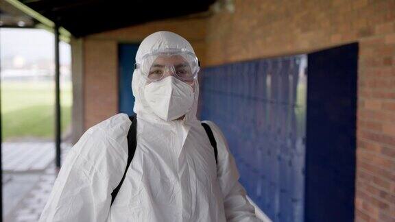 一名年轻人正在为学校走廊消毒他戴着生物安全防护椅、护目镜和防护面罩然后面对摄像头