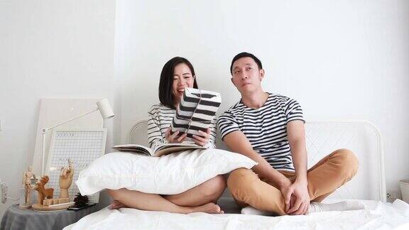 亚洲女人正在阅读杂志与惊喜礼盒与她的男友在他们的卧室休闲活动的惊喜礼盒与他的男友