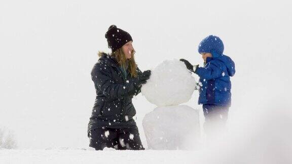 3岁的白人男孩和他的白人母亲在她30多岁(都穿着冬天的衣服)一起堆雪人在一个下雪阴天