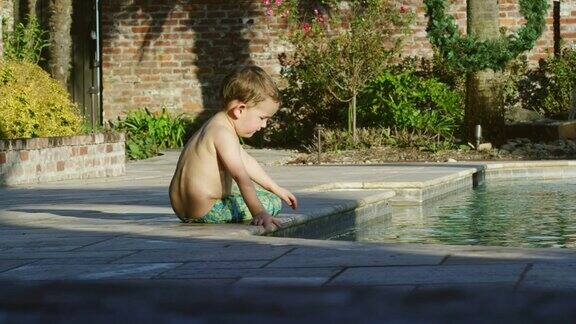 一个阳光明媚的夏日一个三岁的白人男孩爬到游泳池边把手伸进水里