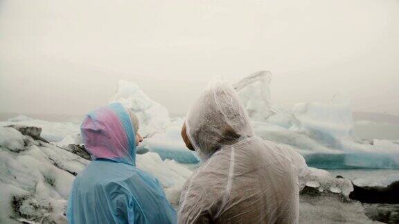 后视图的年轻夫妇在雨衣站在冰泻湖在冰岛和看冰川与灰烬著名的景象