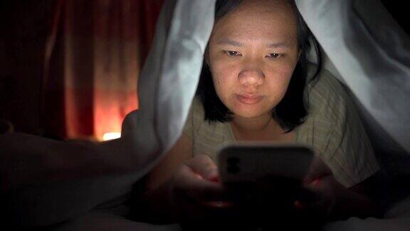 一名亚洲女性晚上在卧室的白毯子下使用智能手机