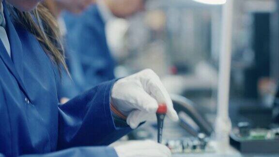 一个女电子工厂工人在蓝色工作服和防护眼镜组装智能手机的特写镜头高科技工厂设施与更多的员工在后台