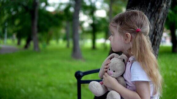 在一个阴天里一个小女孩带着一只玩具兔子坐在公园的长椅上