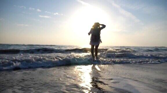 年轻快乐的女人穿着衬衫和背包跑到海边用她的脚泼水在日出漂亮的女孩戴着帽子享受暑假或假期旅行后视图慢动作