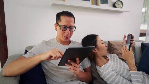 30多岁的亚洲青年男女坐在舒适的客厅沙发上玩手机幸福的一对网民穿着休闲衣服躺在沙发上亚洲的技术使用概念