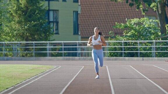 快乐和微笑的健身妇女在浅蓝色运动上衣和紧身裤在一个体育场慢跑她在一个温暖的夏日午后跑步运动员在跑道上进行例行运动练习