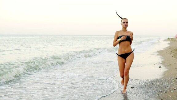 年轻迷人的女人在黑色皮革泳衣在海边的海滩上跑步当女人在锻炼时她的马尾辫在风中摆动Slowmotion拍摄