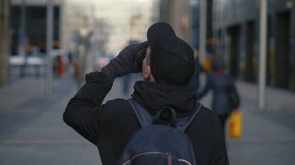 一个戴着黑色帽子背着背包的男人走在城市街道上喝着热咖啡