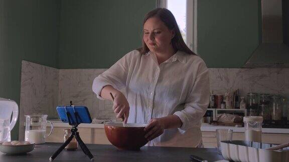 快乐的超重女面包师在厨房里做面团按照食谱把手机放在三脚架上